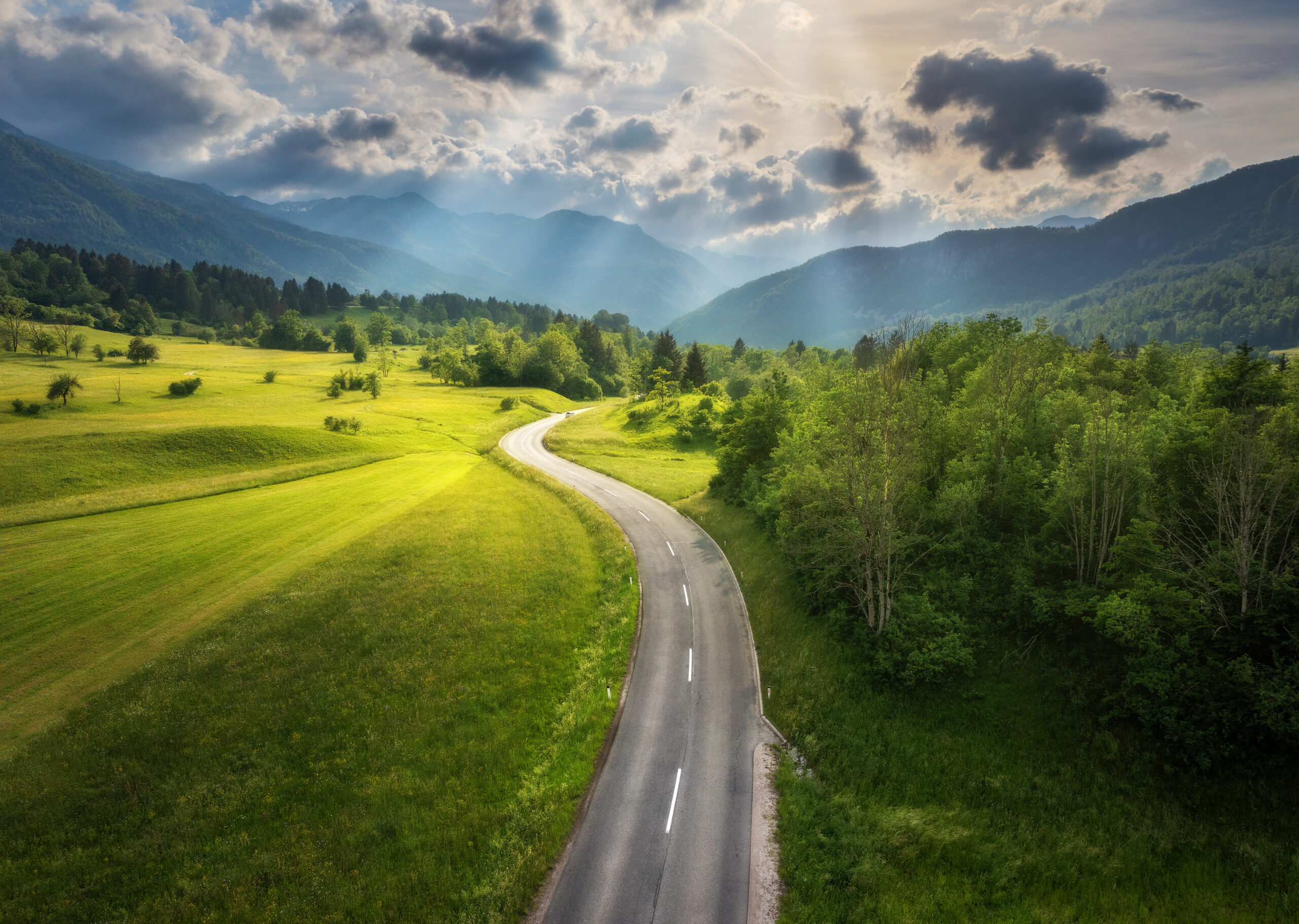 Aerial view of road in green meadows at sunset in summer. Top view from drone of rural road, mountains, forest. Beautiful landscape with roadway, sun rays, trees, hills, green grass, clouds.